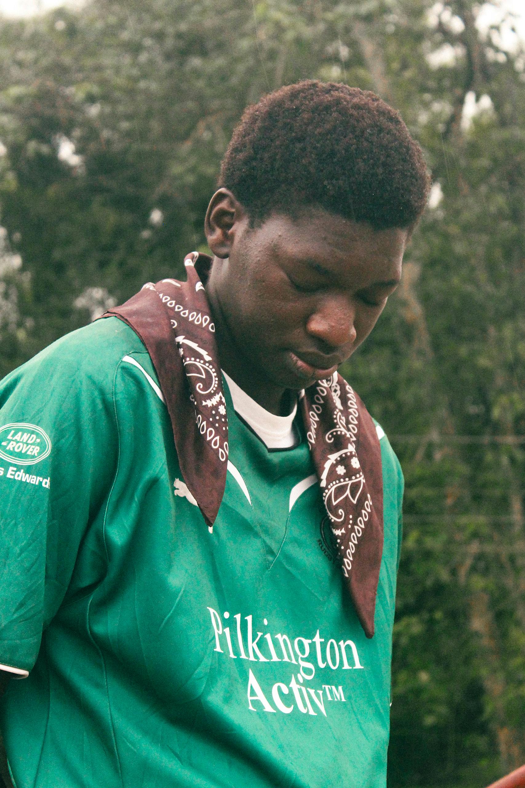 A young male soccer player wearing a green jersey stands outdoors, looking thoughtful.