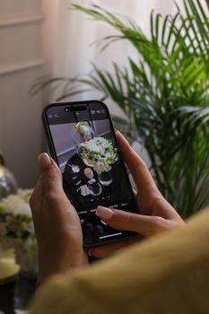 A close-up of a person photographing a white floral bouquet indoors next to a lush green plant.
