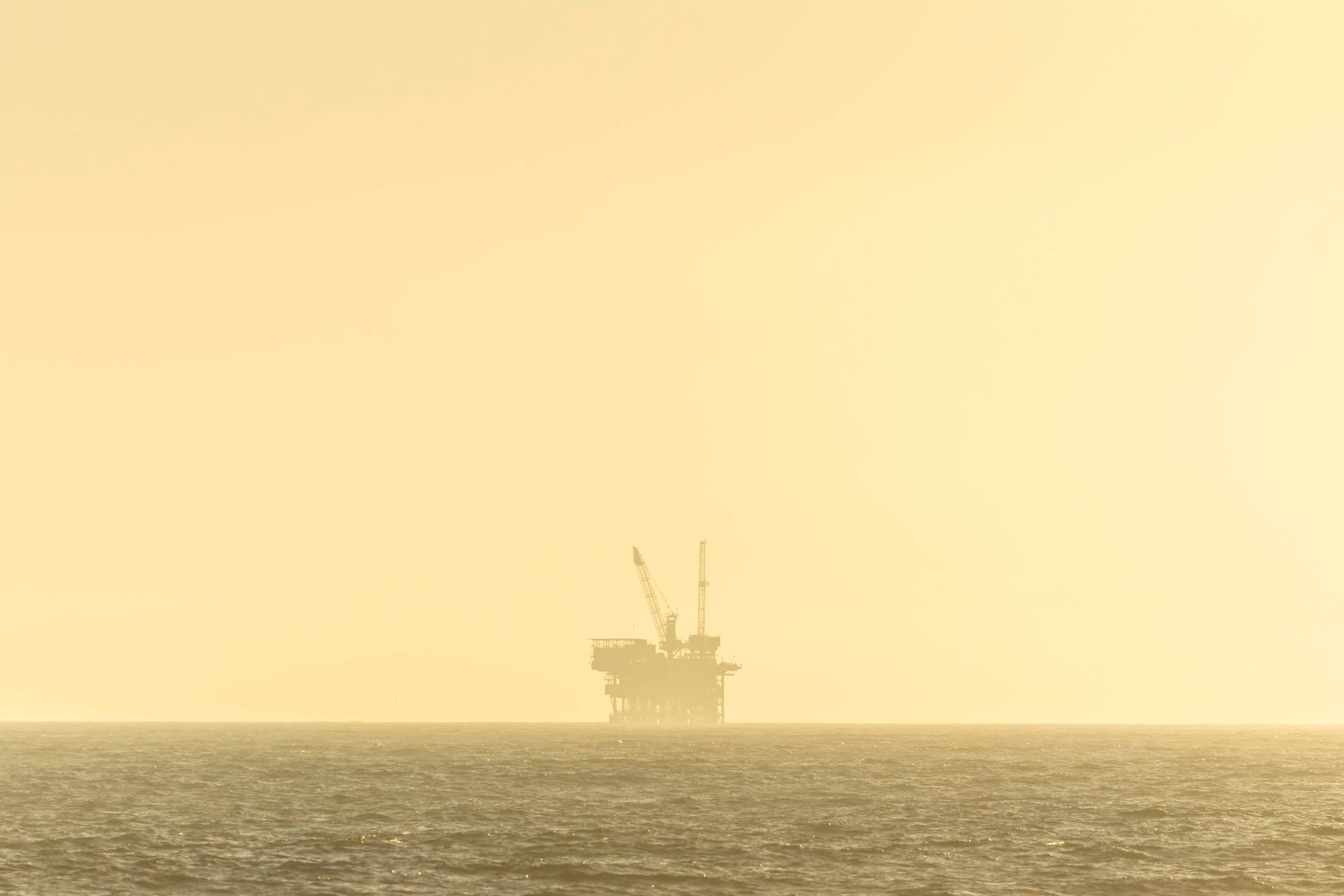 Distant view of an offshore oil platform against a golden sky at sunset over a calm sea.