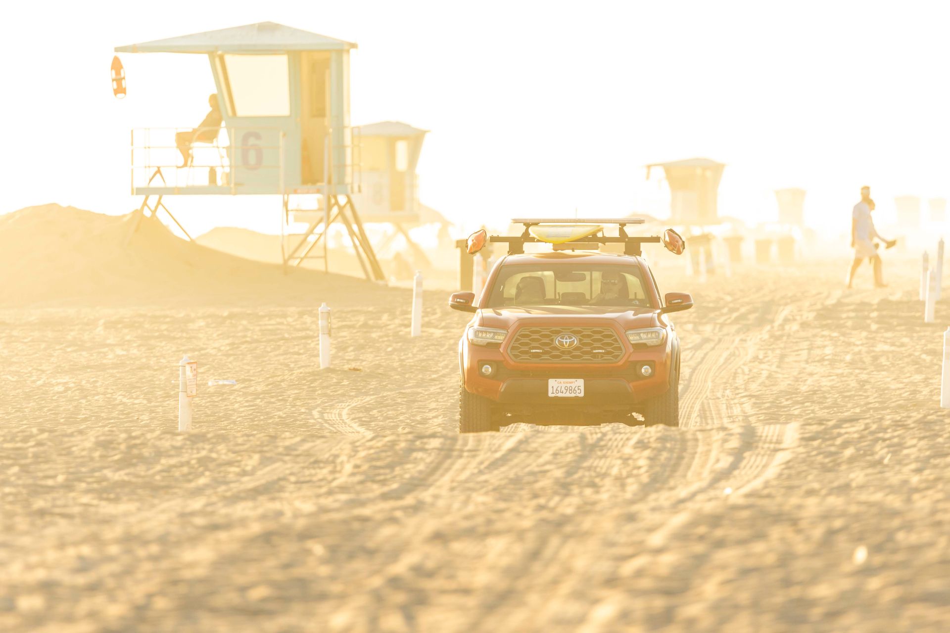 A red lifeguard truck drives on a sunlit California beach with visible lifeguard towers.