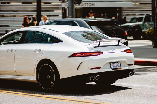 A sleek white luxury car cruising in an urban setting with people in the background on a sunny day.