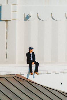 A man wearing casual attire sits alone on an urban building ledge in contemplative solitude.
