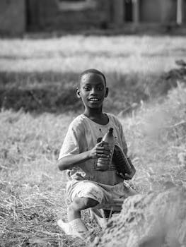 Black and white portrait of a smiling child holding bottles, seated outdoors in Burundi.
