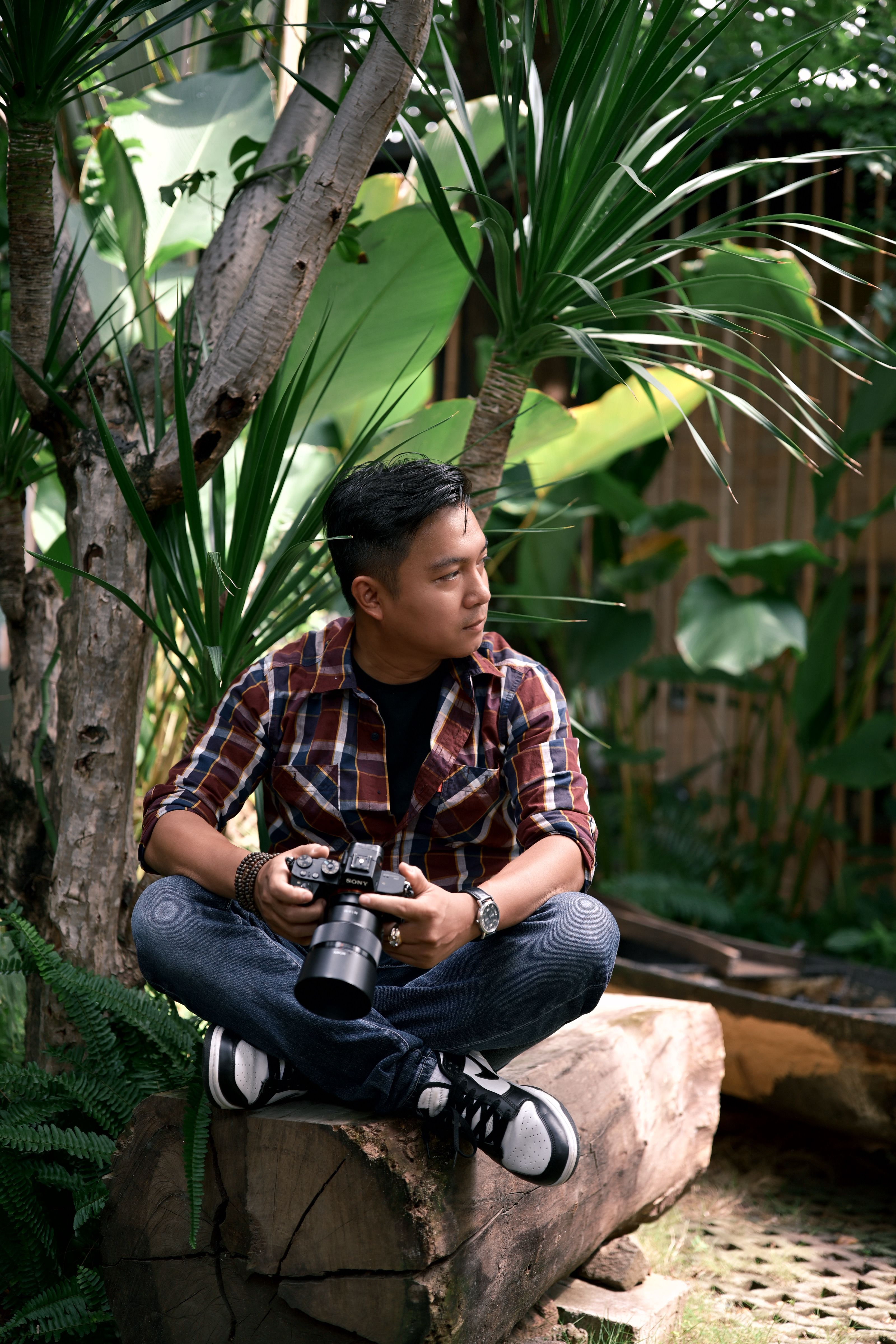 Man holding camera, seated on log in lush tropical garden, wearing plaid shirt.