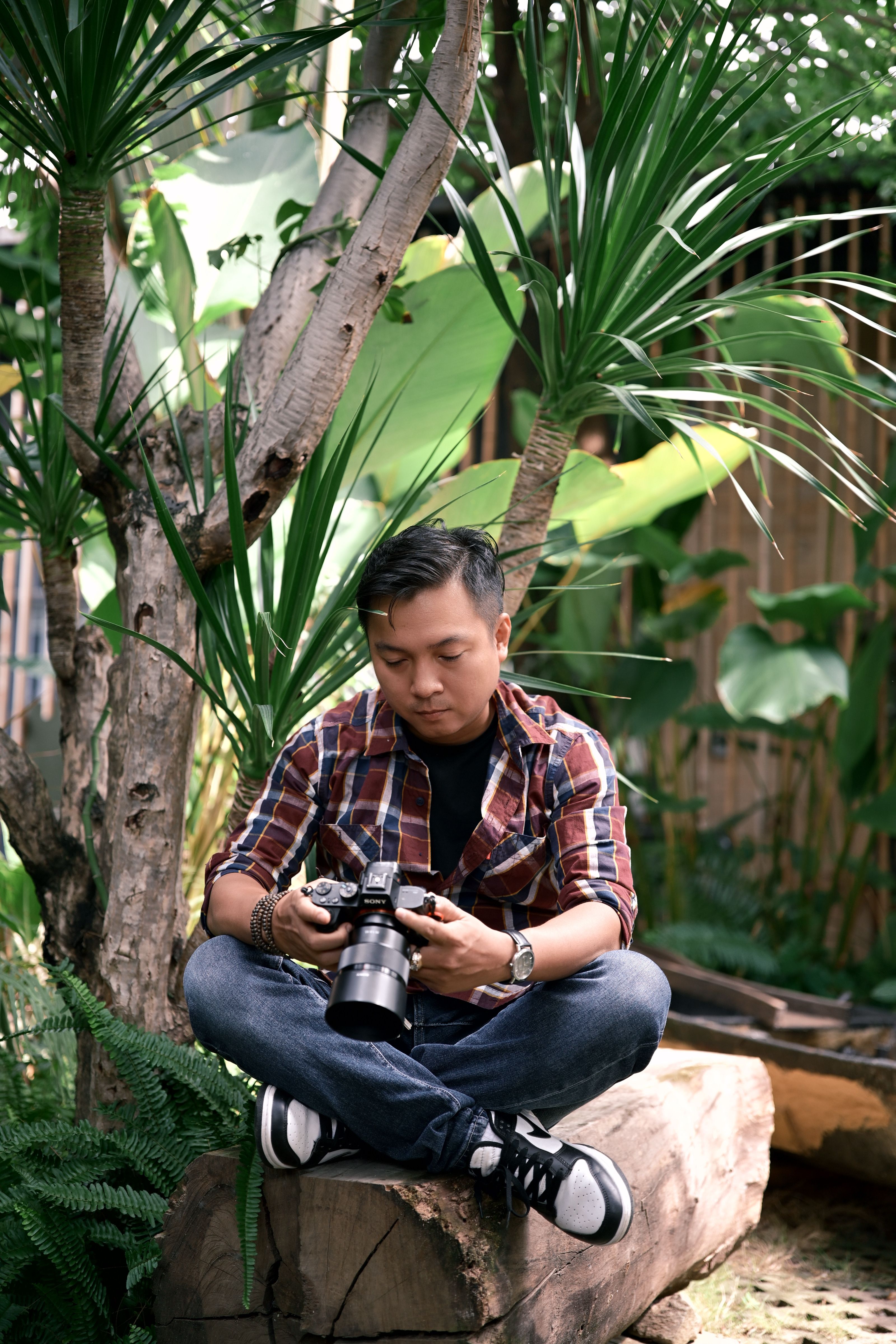Young male photographer sitting on a log with a camera in a vibrant green garden.