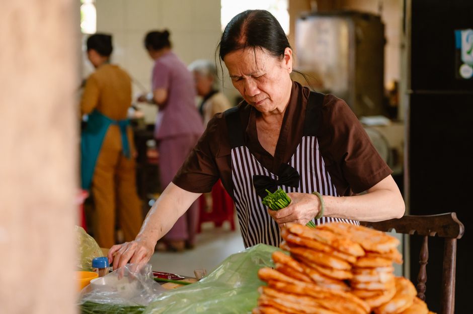 An elderly woman wearing an apron prepares vegetables in a bustling indoor kitchen setting.
