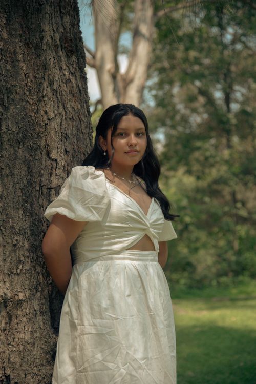 Free A young woman in a white dress stands by a tree in a serene outdoor setting, embodying grace and tranquility. Stock Photo