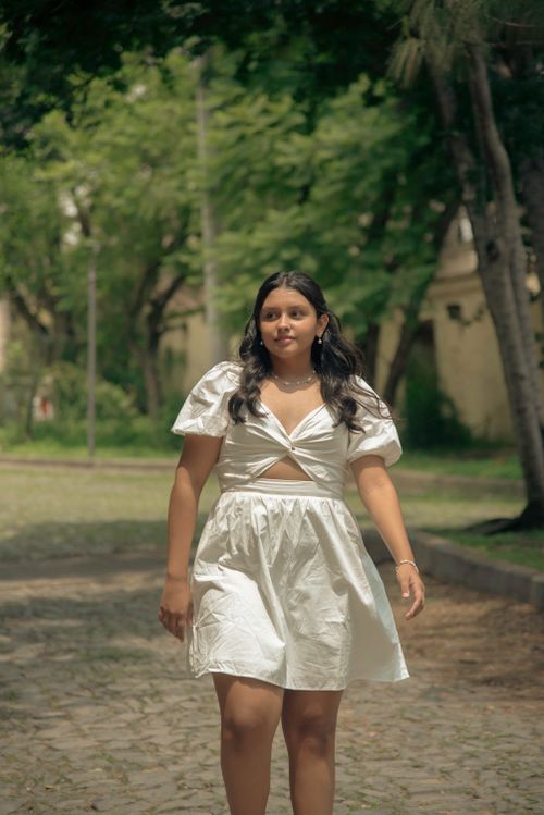 Free A young woman wearing a white dress strolls through a lush green park on a sunny day. Stock Photo