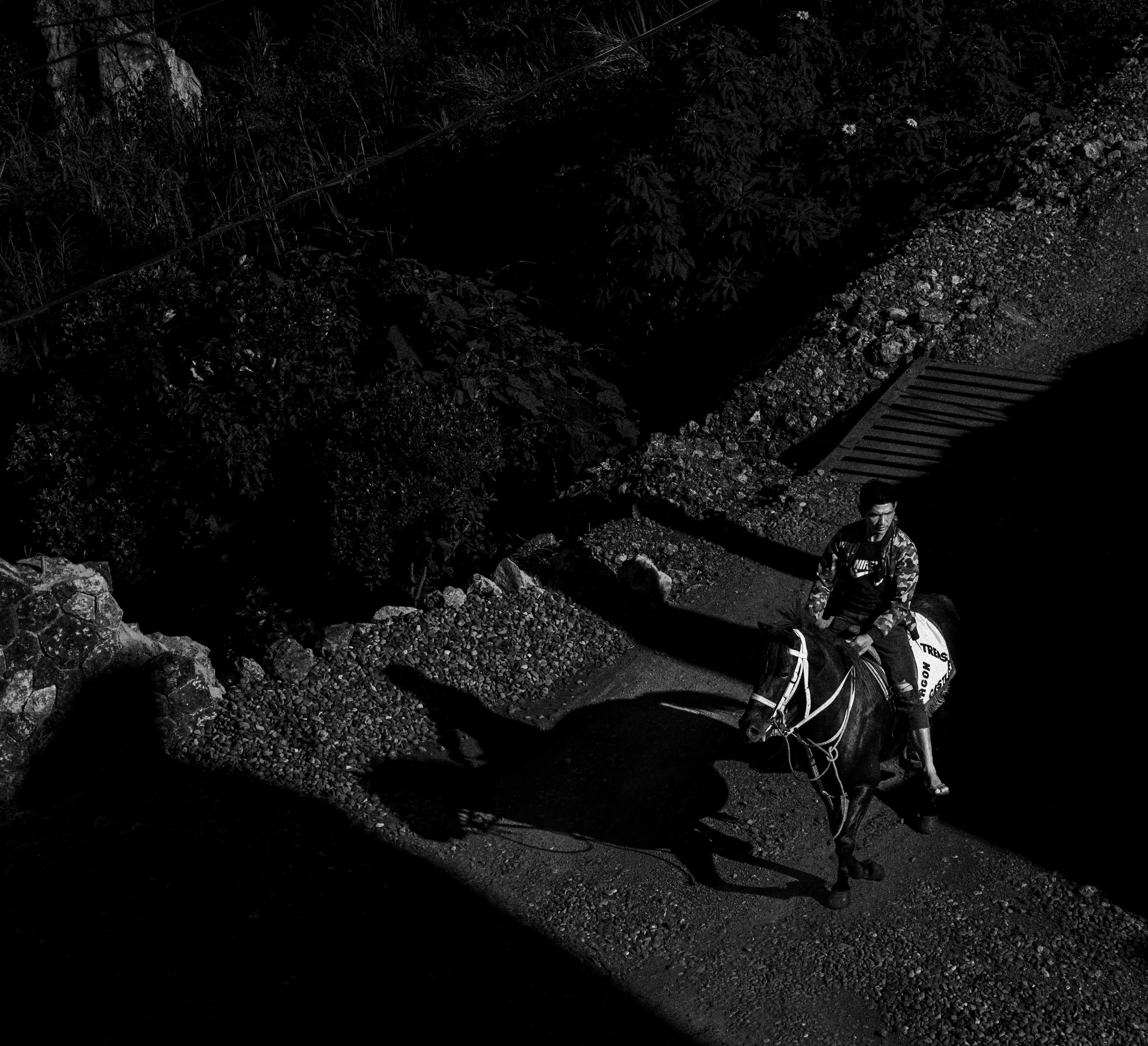 Black and white aerial shot of a man riding a horse on a rural dirt road, casting strong shadows.