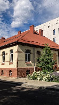 A picturesque brick building with a red tiled roof, surrounded by lush greenery under a blue sky.