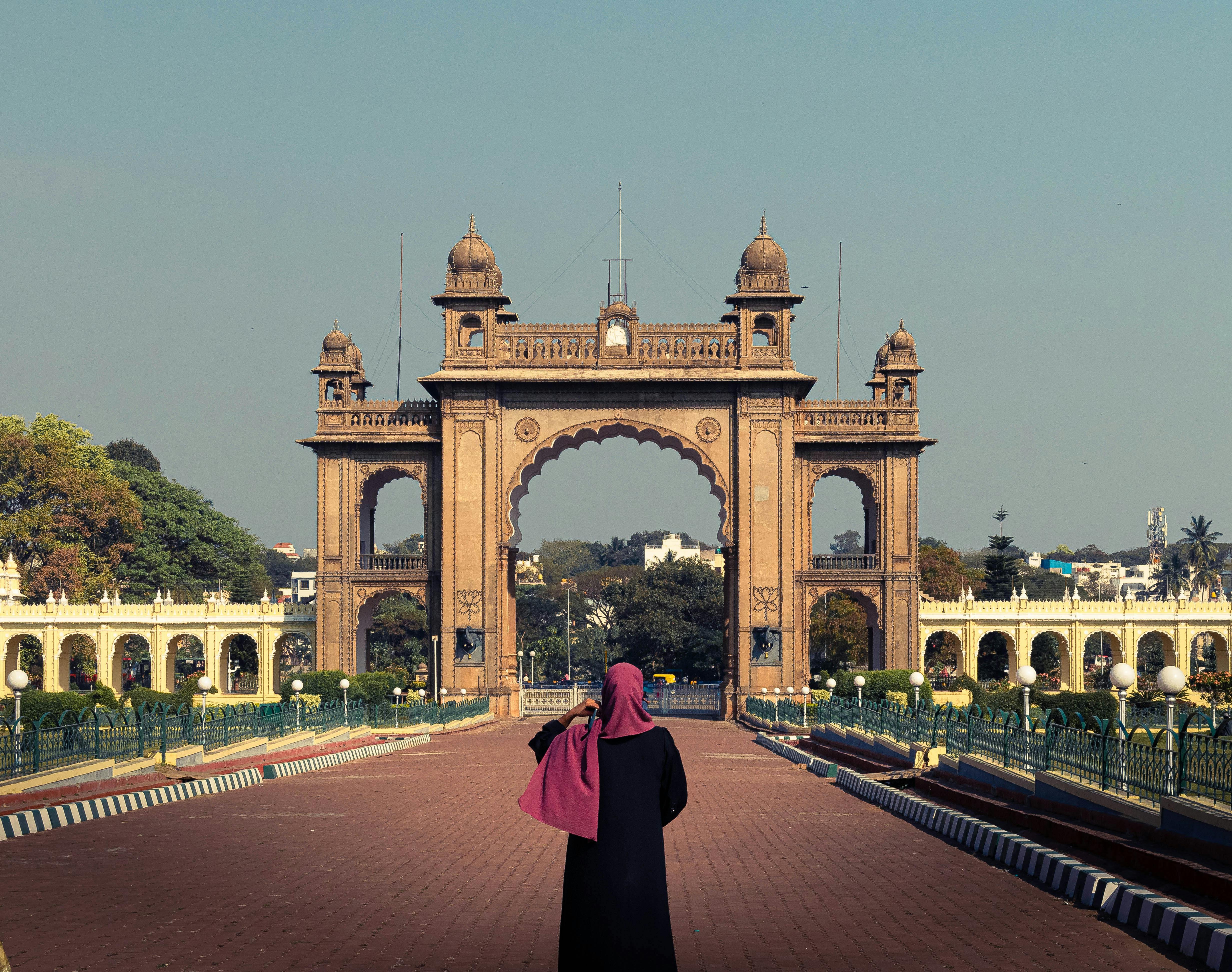 A woman in a black hijab photographs the historic Mysore Palace gate on a sunny day.