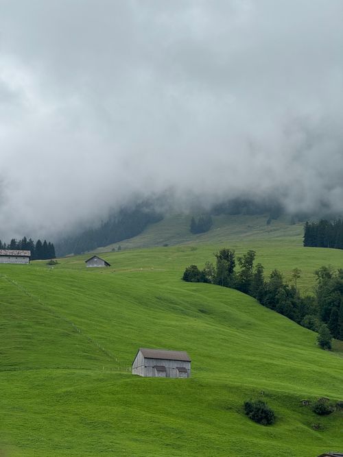 Free Idyllic alpine scene with green hills, rustic barns, and misty clouds. Stock Photo