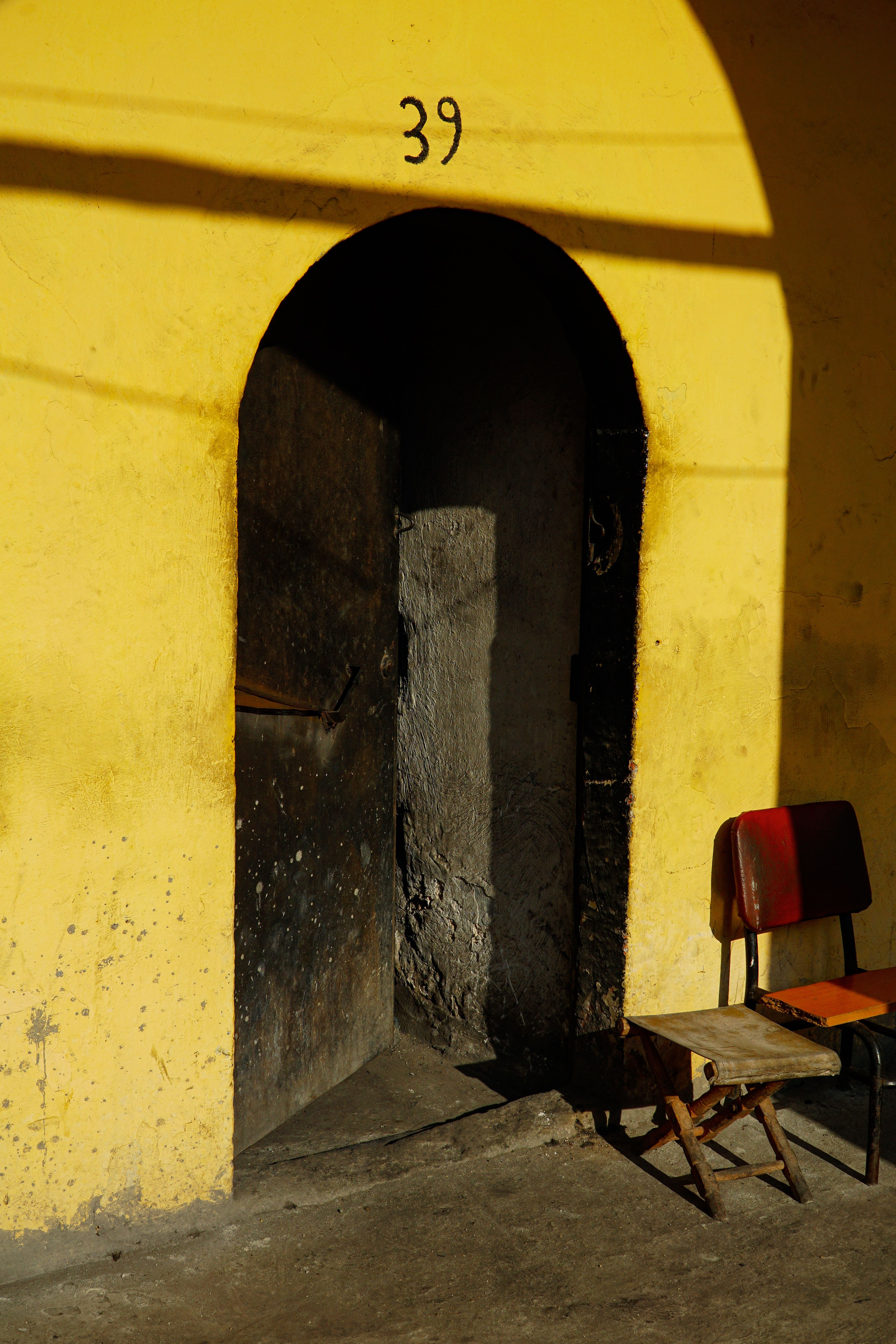 A rustic arched doorway against a vibrant yellow wall, accompanied by a vintage chair casting shadows.