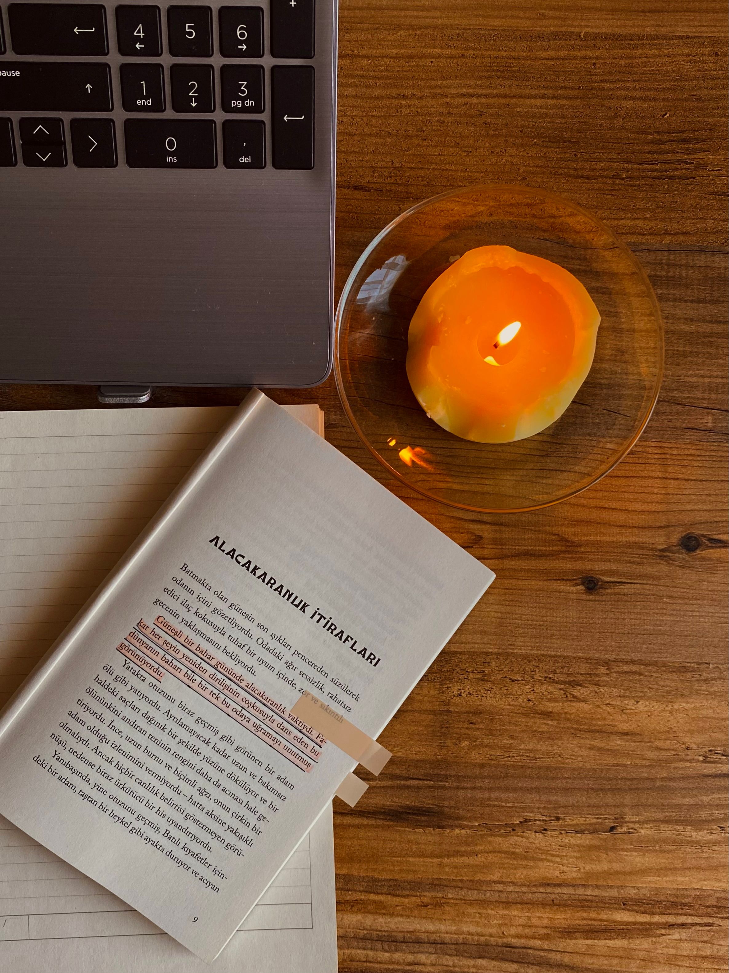 Warm reading setup featuring an open book, lit candle, and laptop on a wooden table.