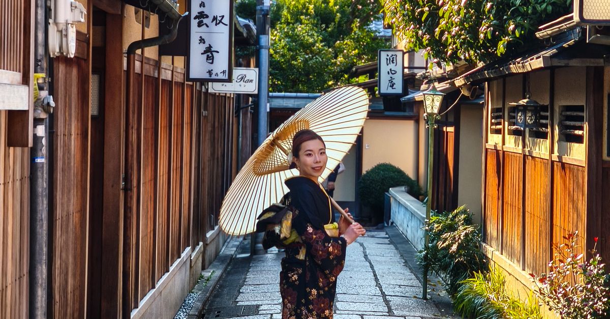 A woman wearing a kimono holds an umbrella in a traditional Kyoto alleyway at sunset.