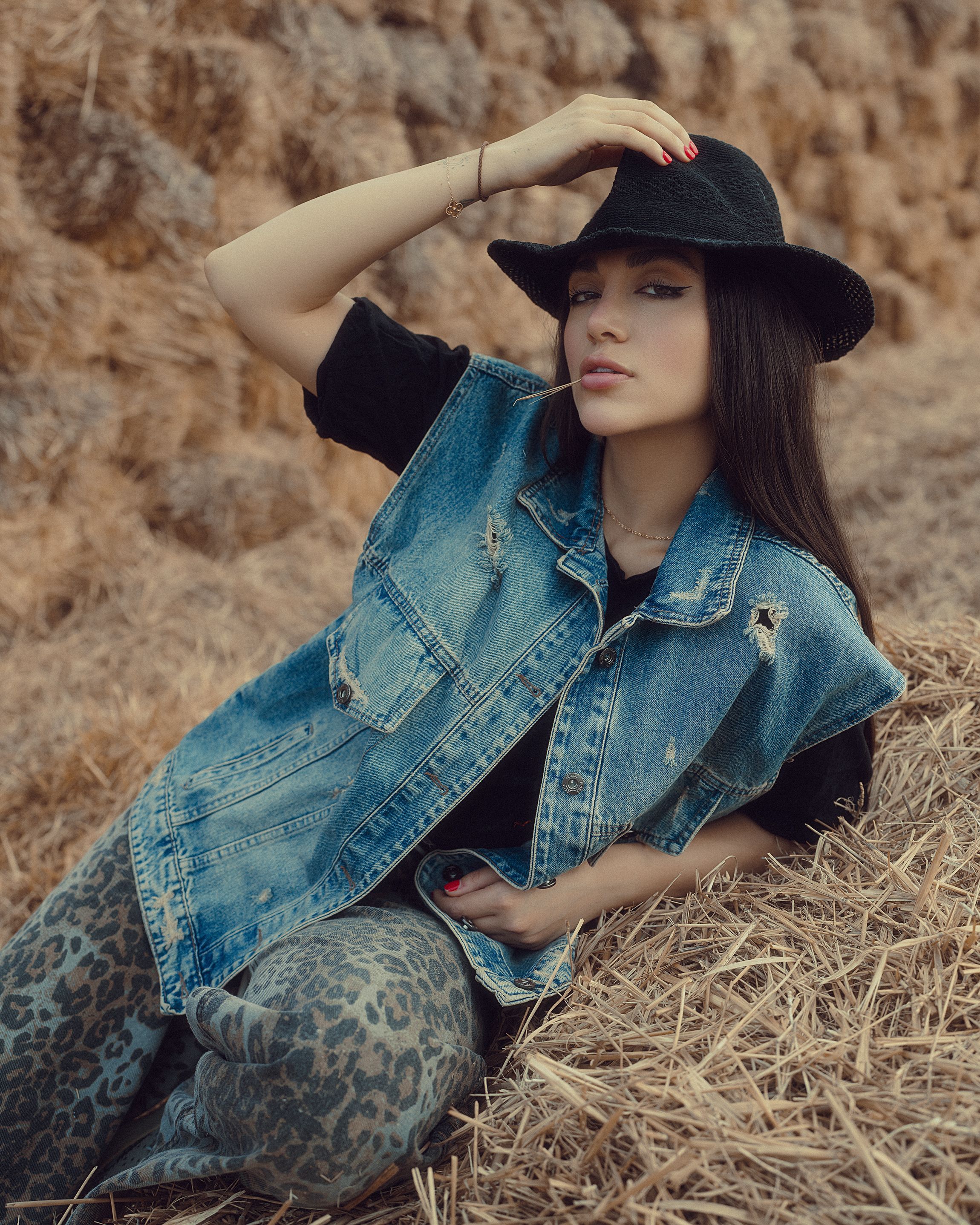 Mulher elegante vestindo um colete jeans e chapéu, sentada em um palheiro ao ar livre. Perfeito para temas de moda e estilo de vida.