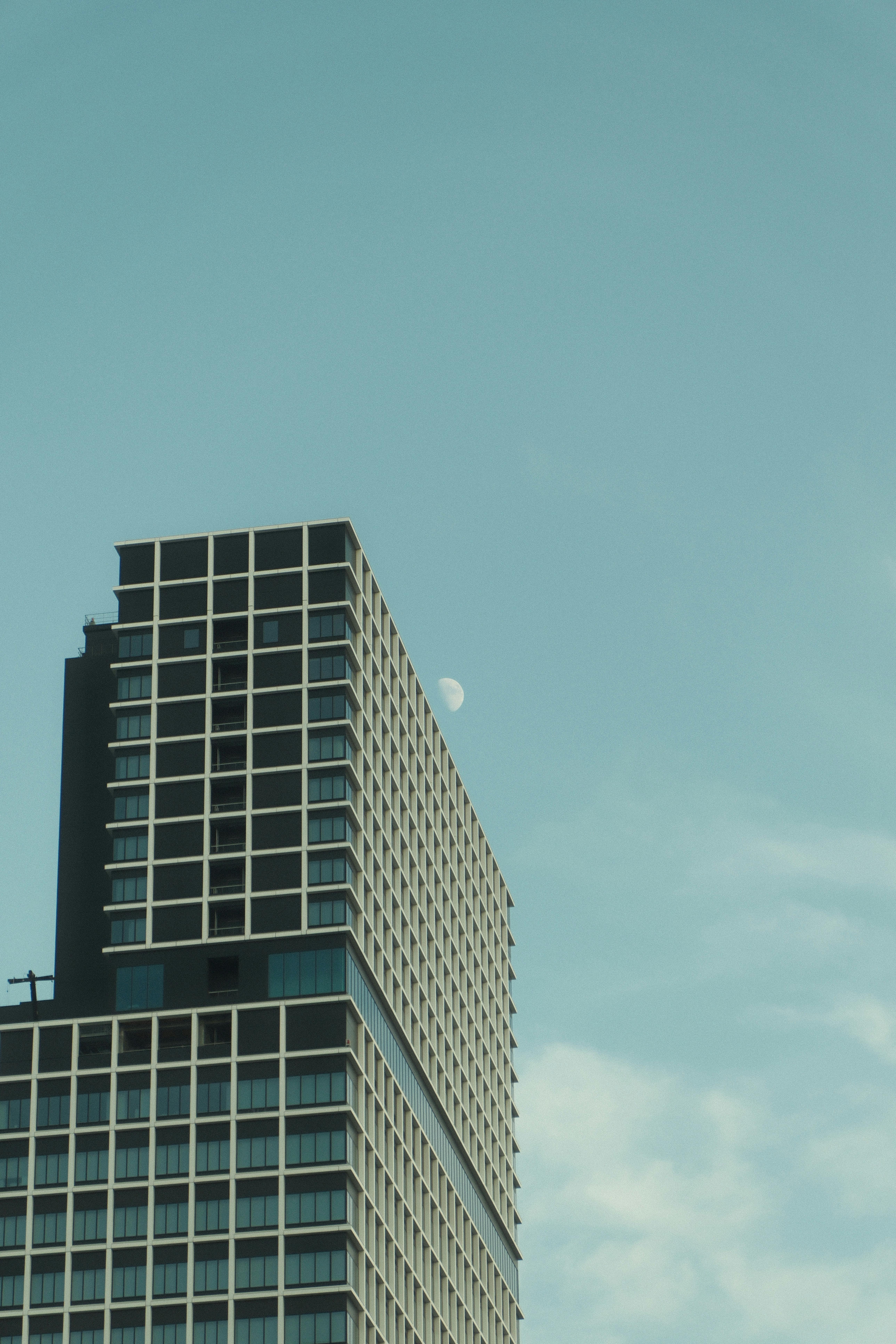 grátis Edifício alto e moderno com a lua contra o céu azul claro, criando um cenário urbano sereno. Foto profissional
