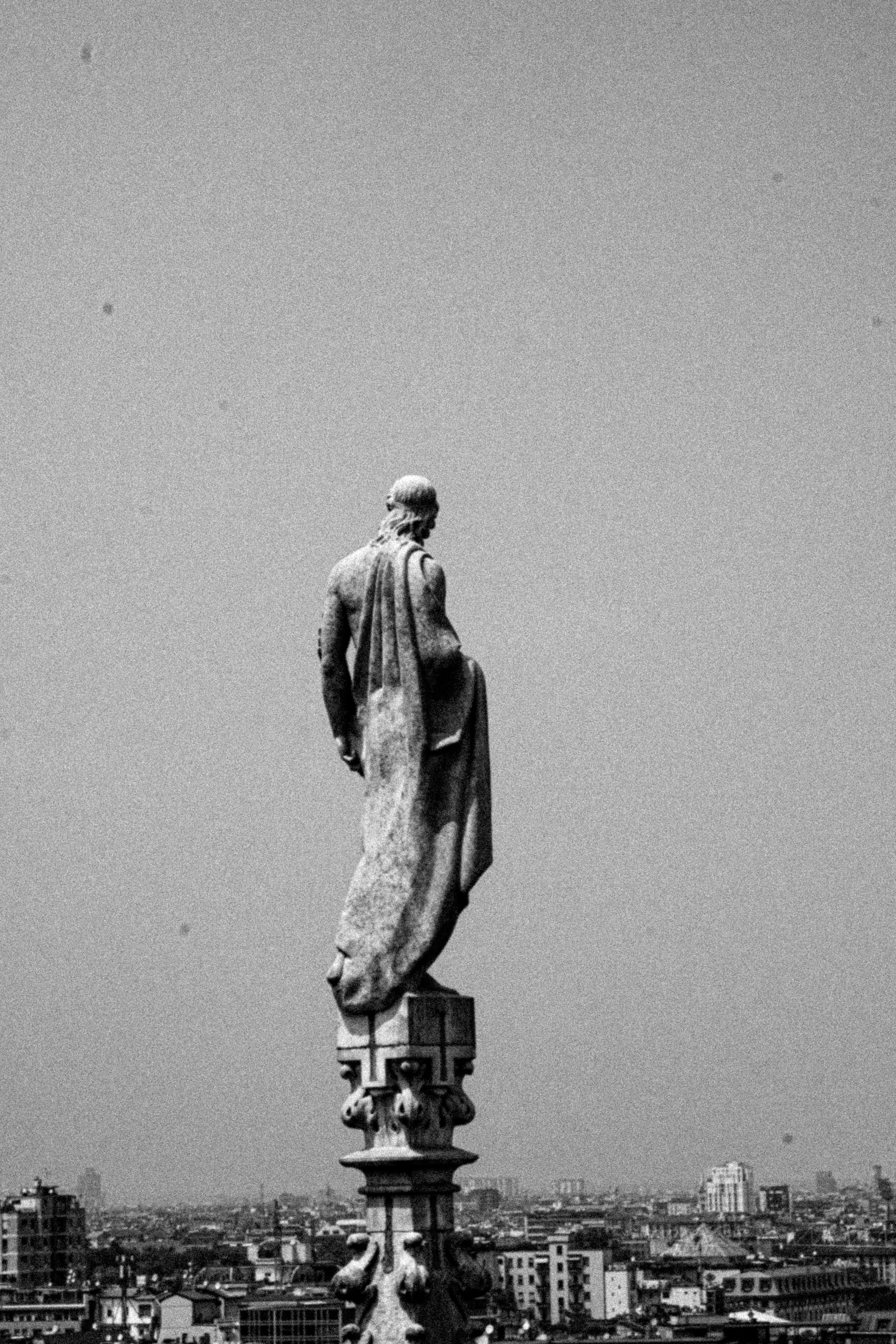 A black and white view of a statue atop Duomo di Milano, overlooking Milan's cityscape.