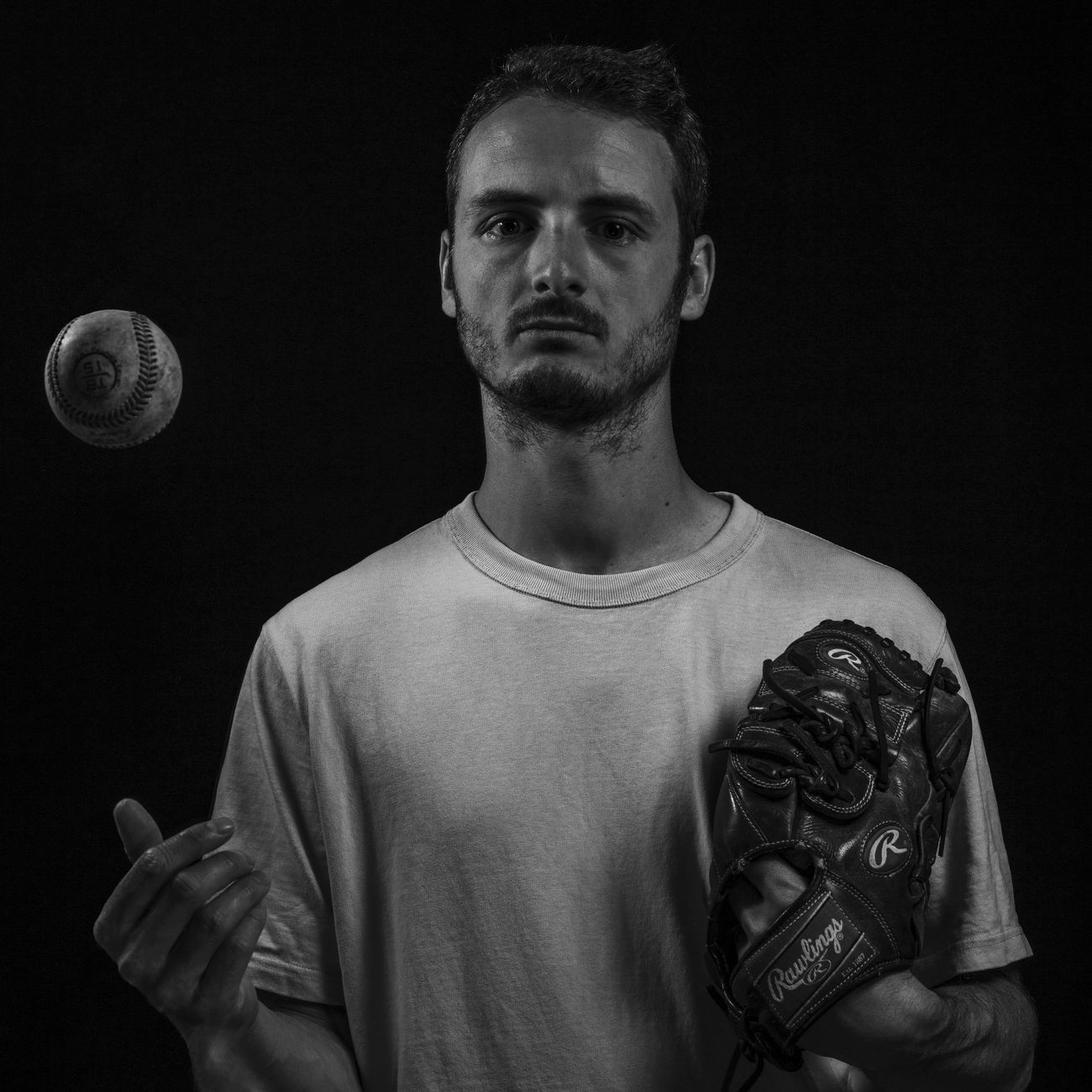 Moody black and white portrait of a man holding a baseball glove and tossing a baseball.