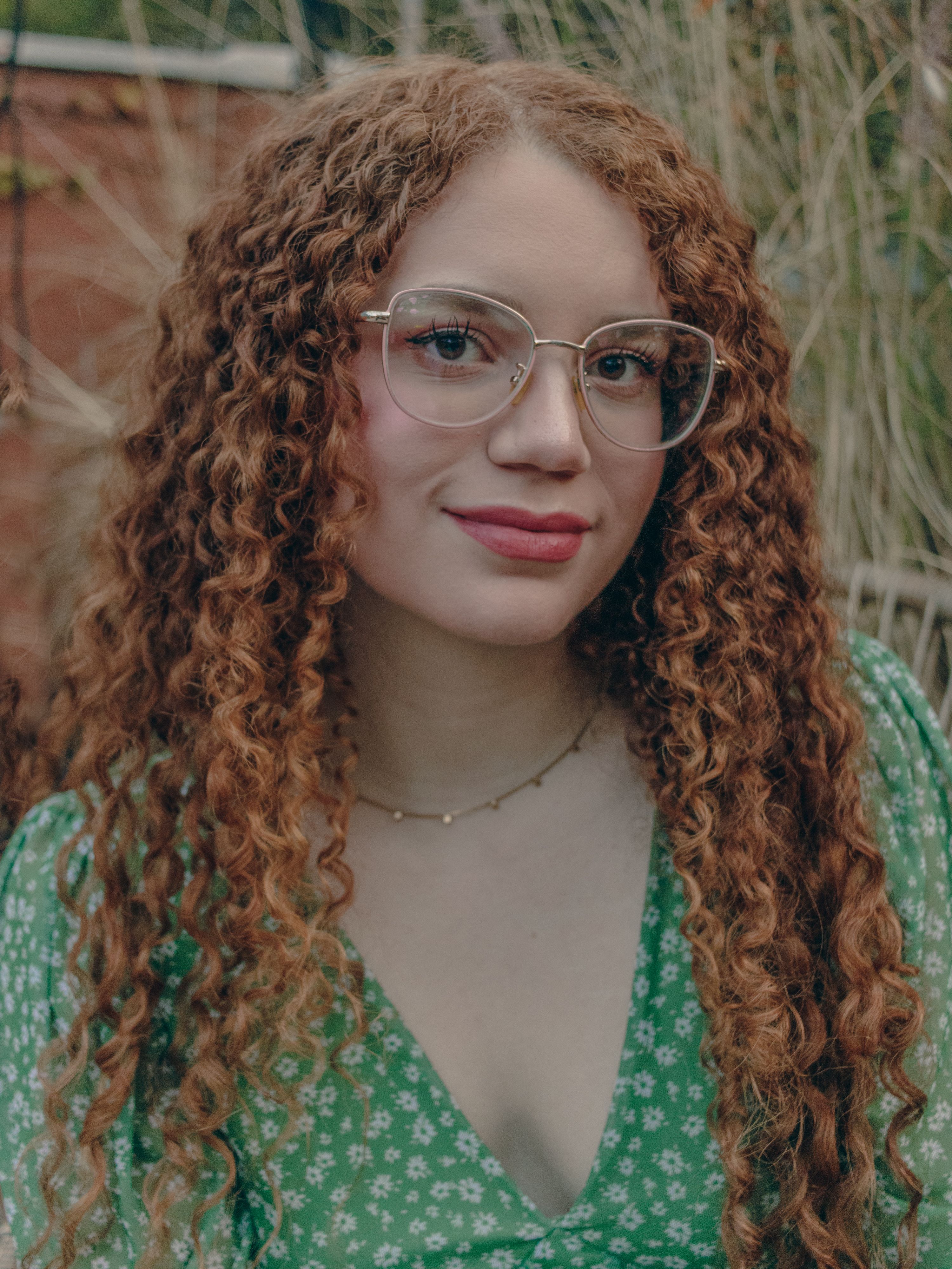Portrait of a young woman with curly hair and glasses wearing a green floral dress.