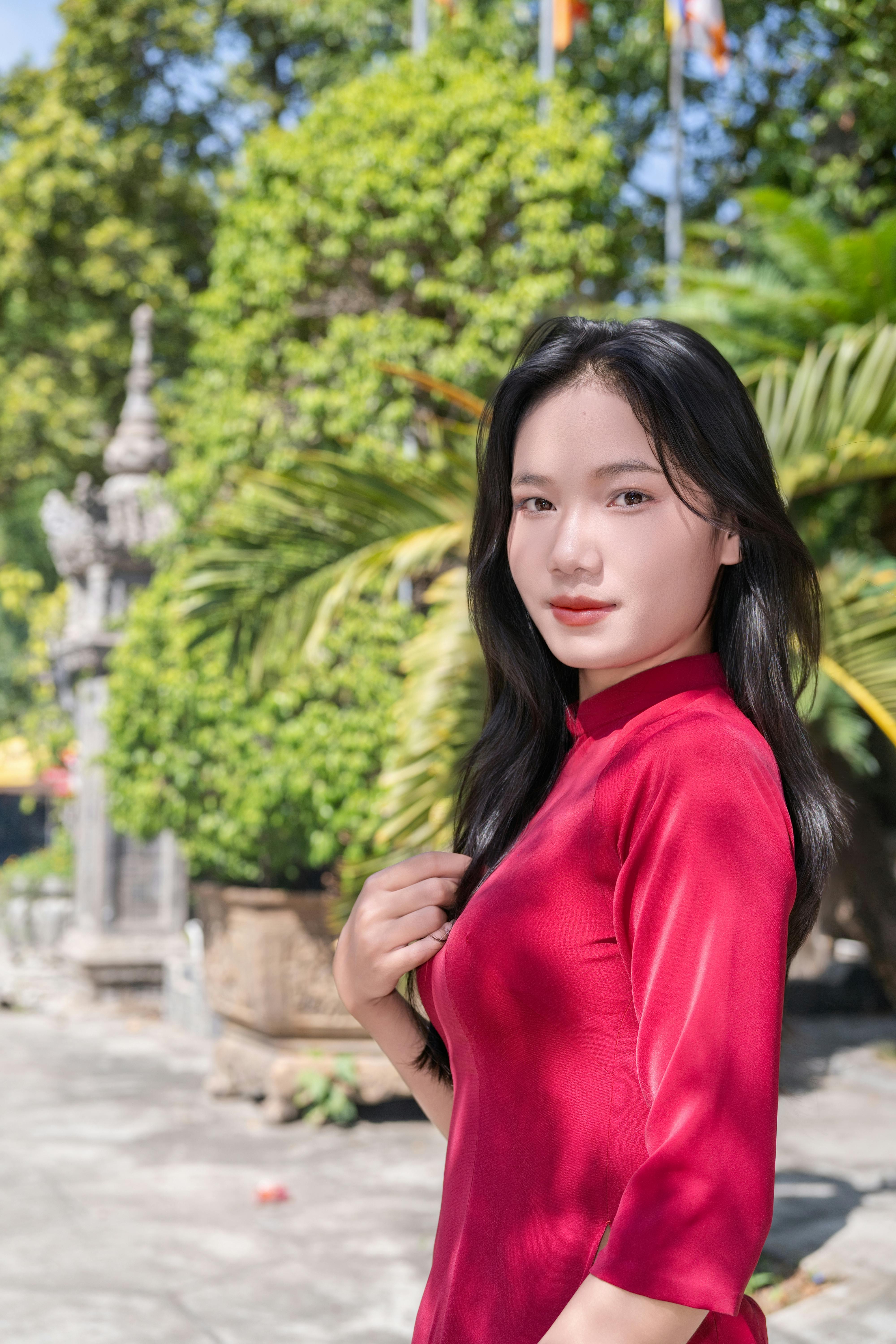Elegant woman in red ao dai posing outdoors on a sunny day with lush greenery.