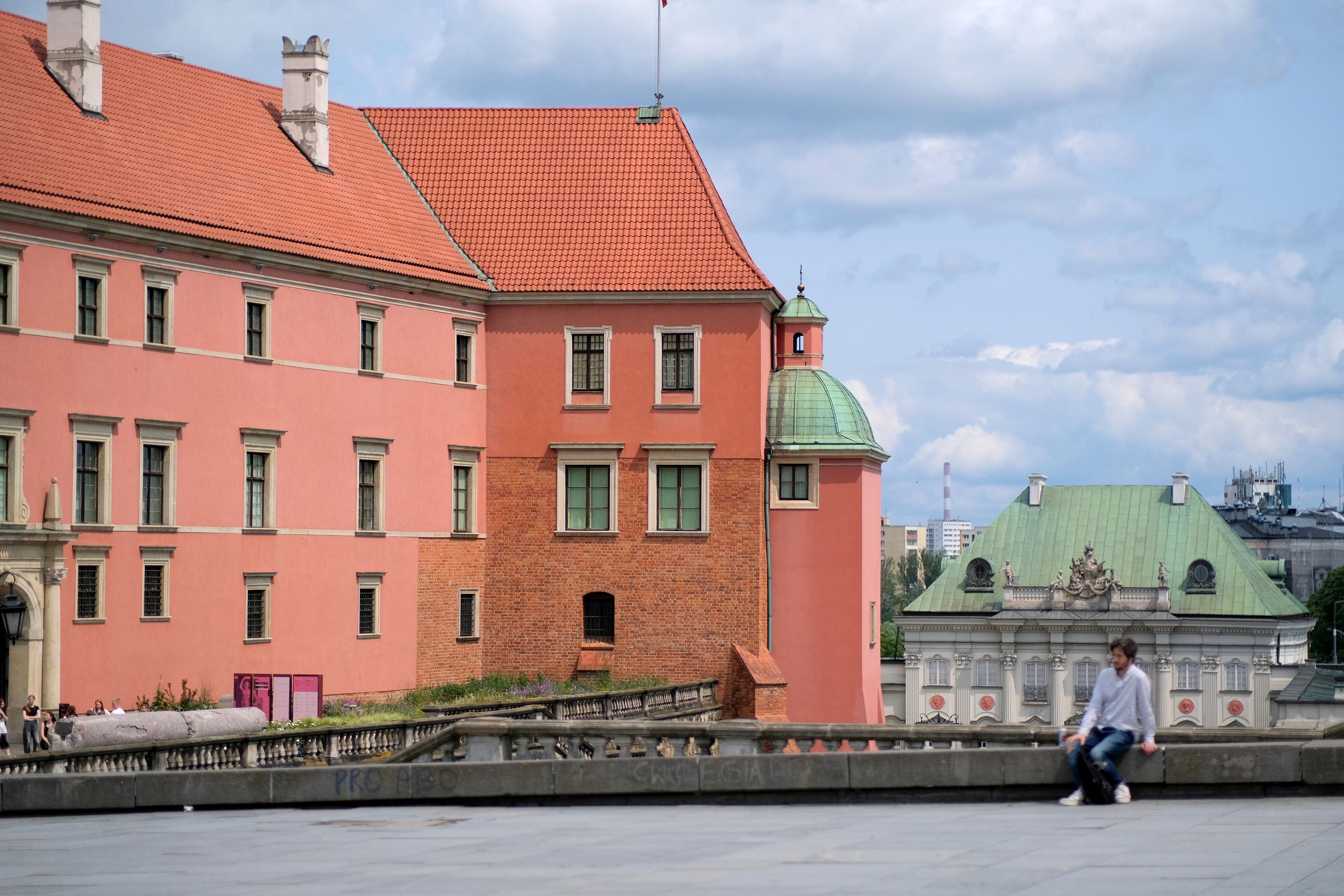 A person sitting near the iconic Warsaw Royal Castle under a bright, partly cloudy sky.