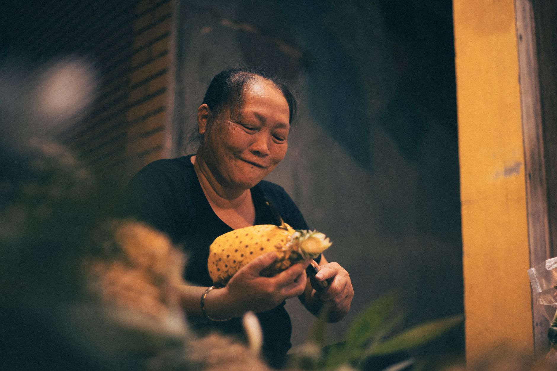 A woman skillfully peels a pineapple at a bustling night market in Hanoi, Vietnam, showcasing urban life.