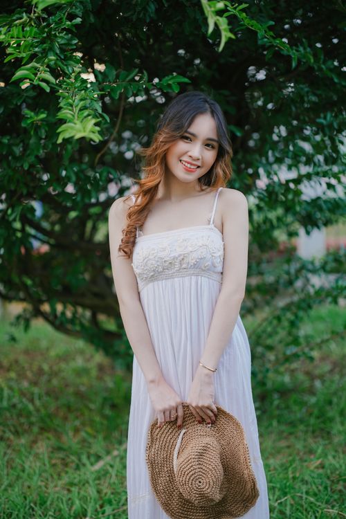Free Young woman in a white dress holding a straw bag, smiling in a lush green outdoor setting. Stock Photo