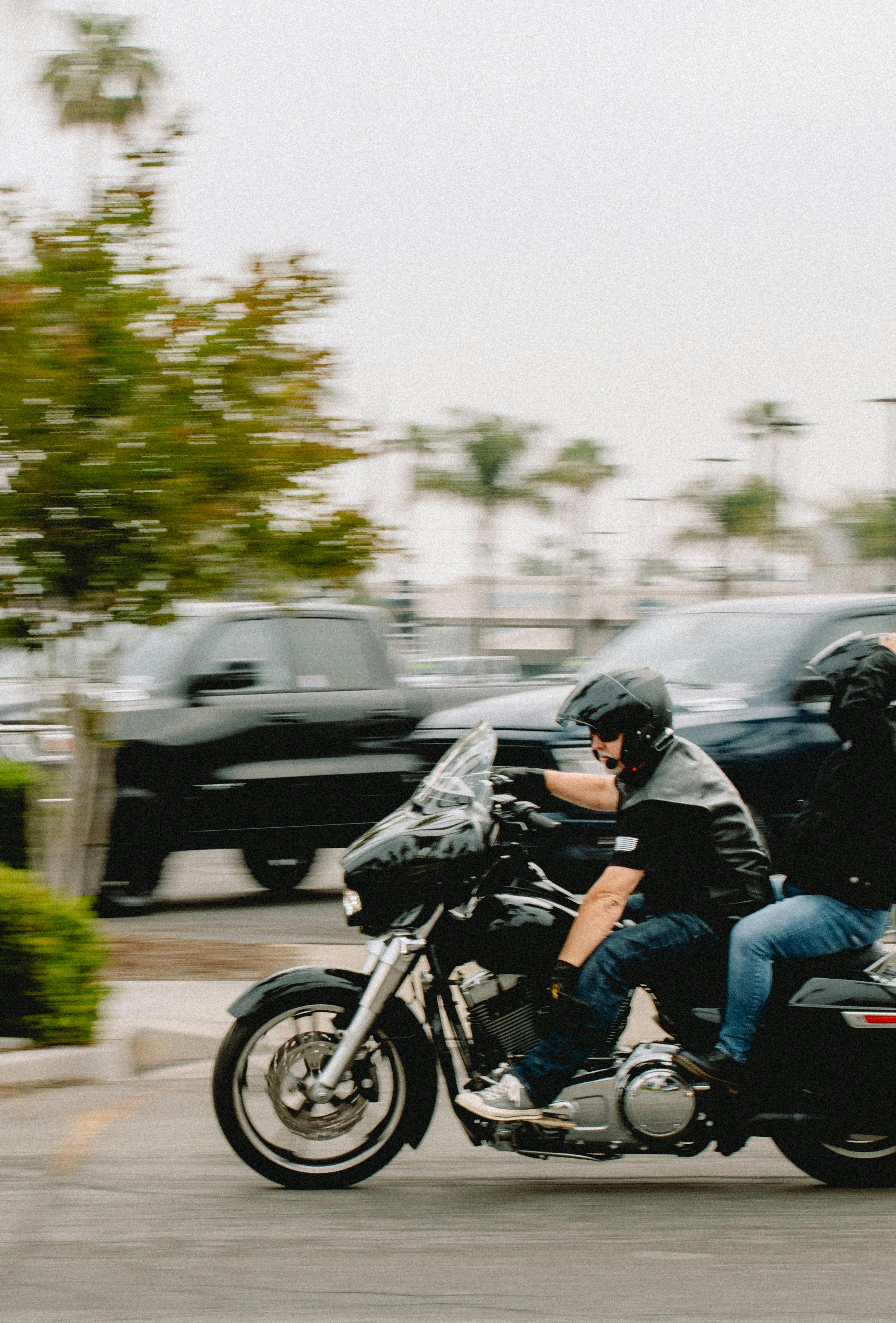 Two bikers riding a Harley-Davidson motorcycle on a street, surrounded by blurred vehicles and trees.