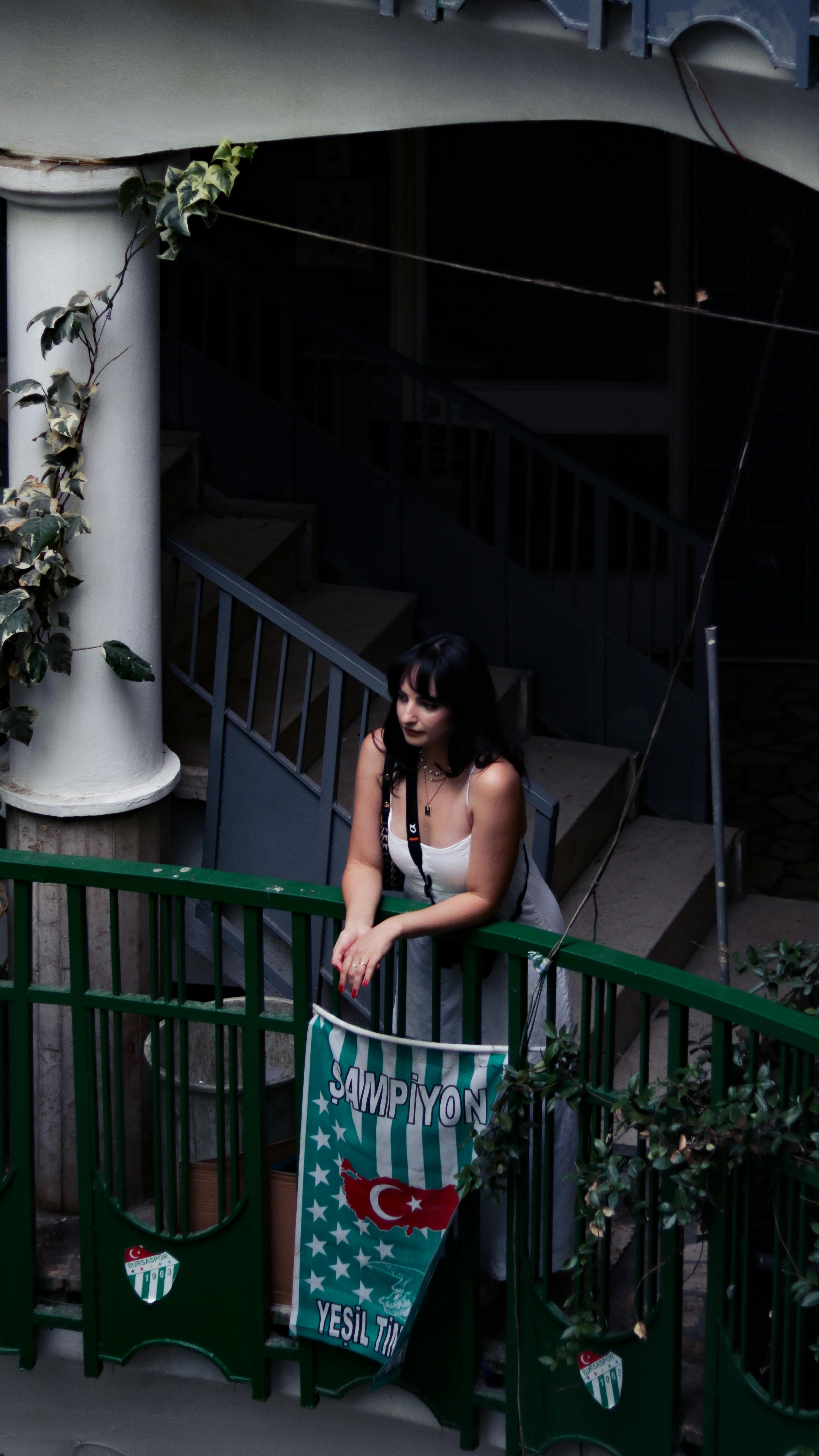 Young woman leaning on railing inside a dimly lit building, featuring a hanging flag.