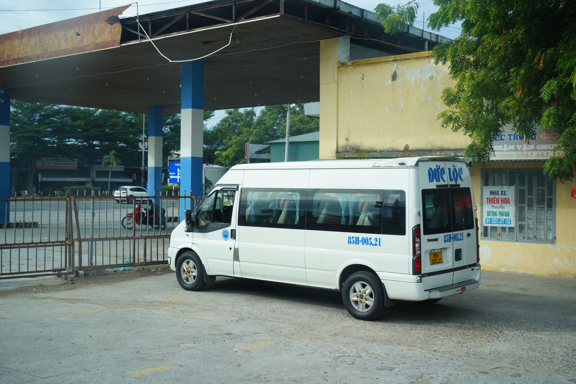 A white minibus is parked at a bus station under a rustic metal canopy.