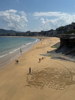 Stunning aerial view of sandy Playa La Concha beach with intricate sand art and distant cityscape in Spain.