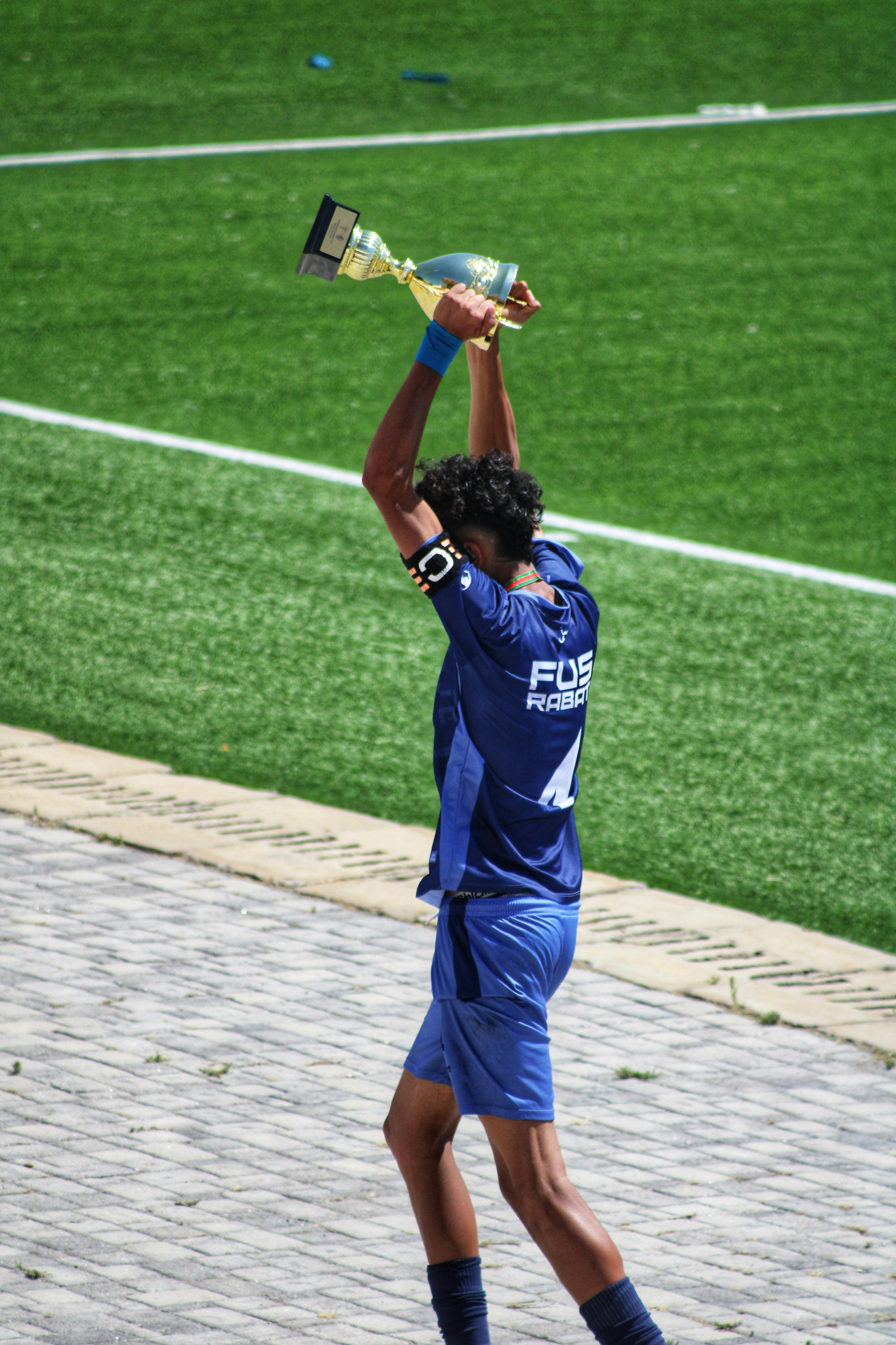 A soccer player in a blue uniform raises a trophy on a sunny field, celebrating victory.