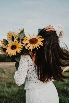 Woman in white dress holding sunflowers in a sunny outdoor field.