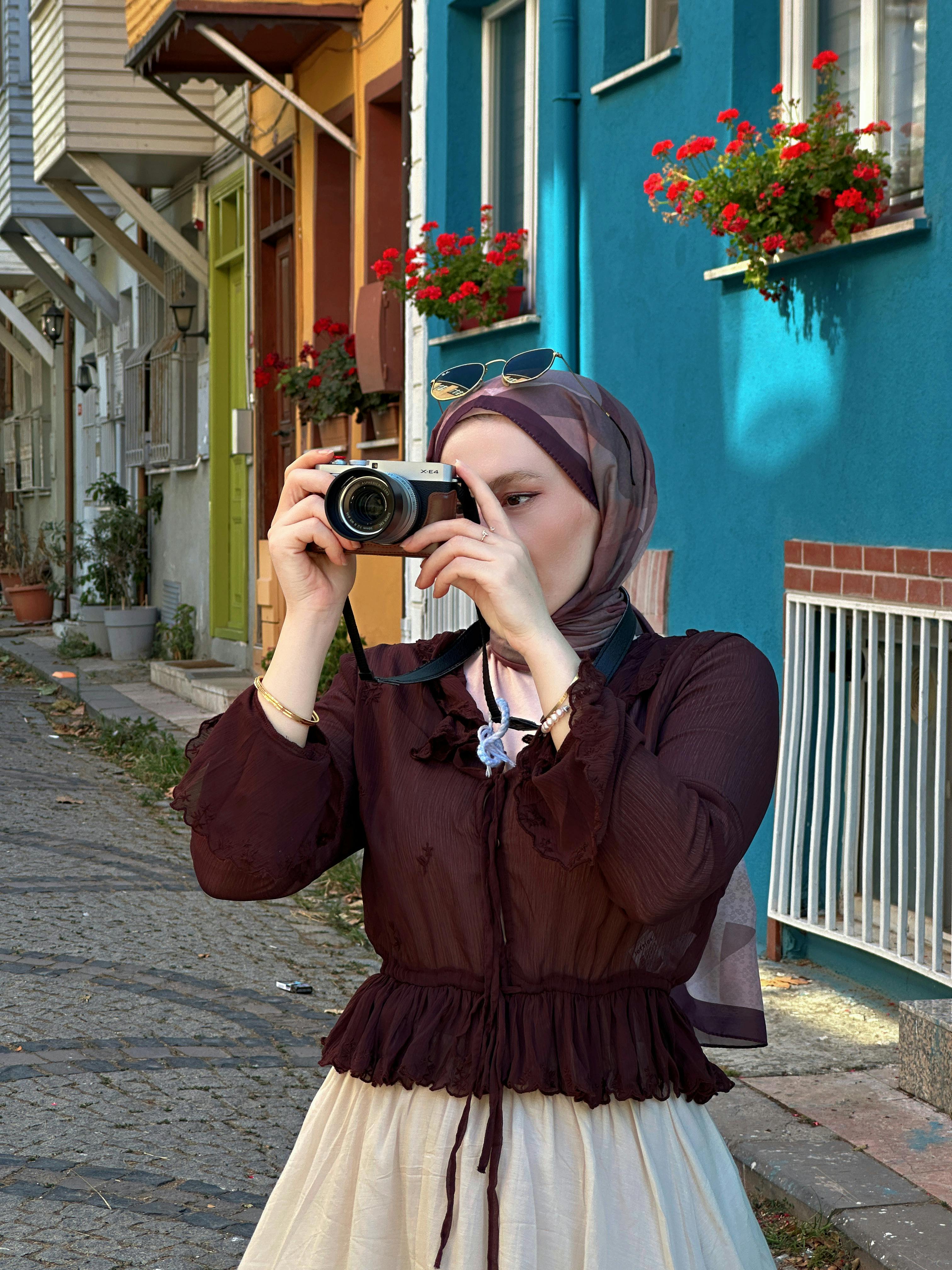 Young woman in hijab photographing on a vibrant, flower-lined street.