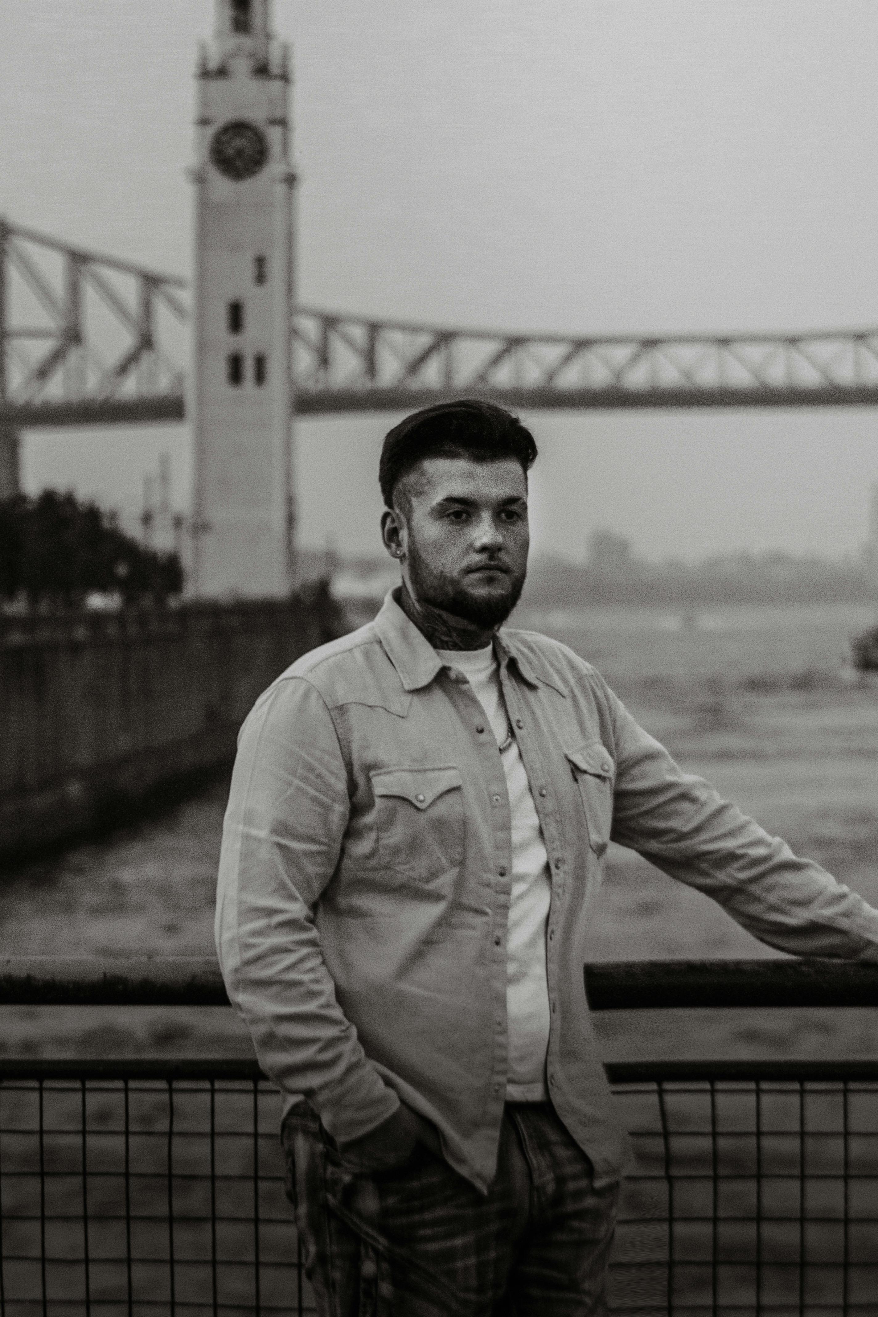 Black and white portrait of a man standing by a river with bridge and clocktower.
