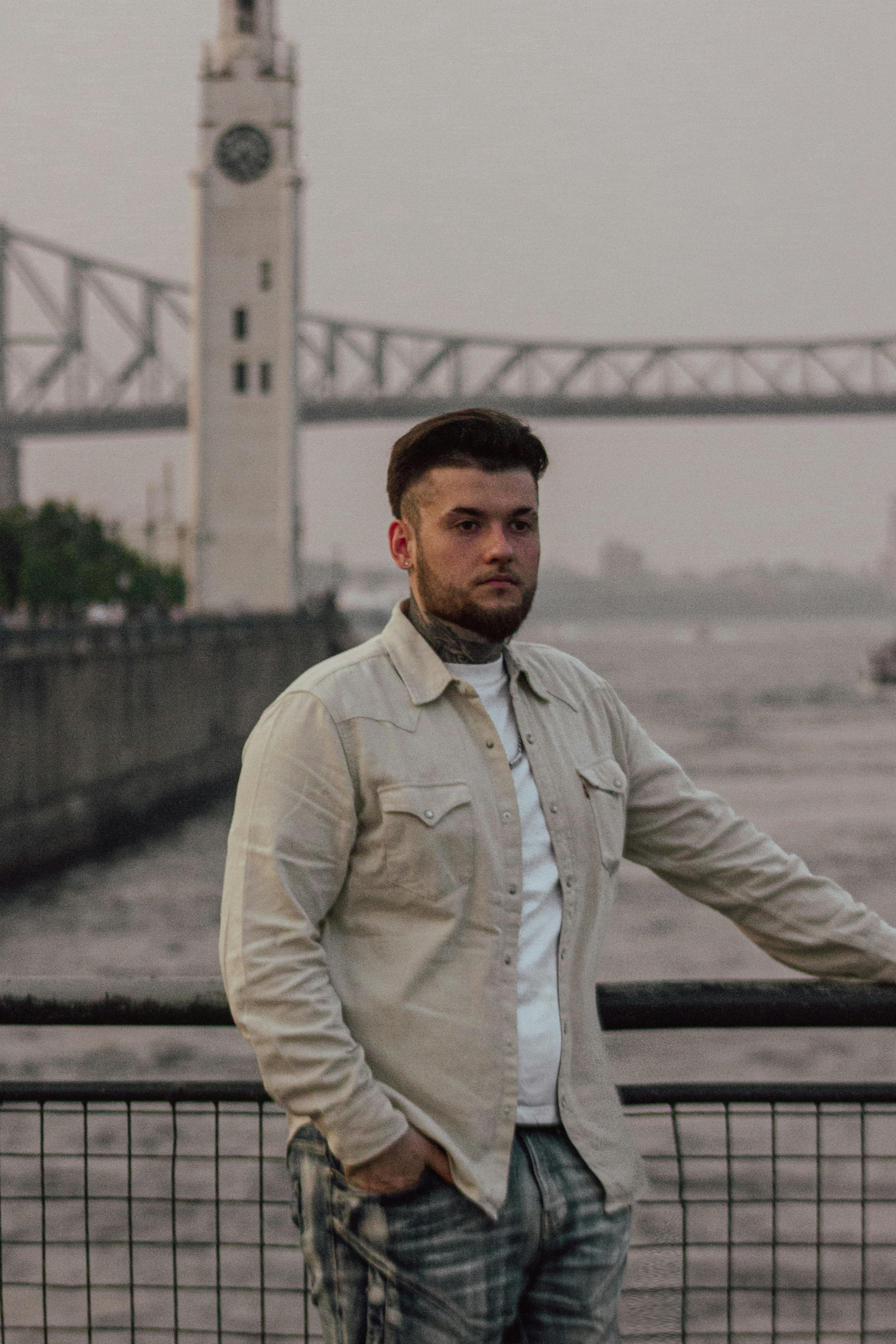 Young man stands by a riverside fence with a clock tower and bridge in the background during twilight.