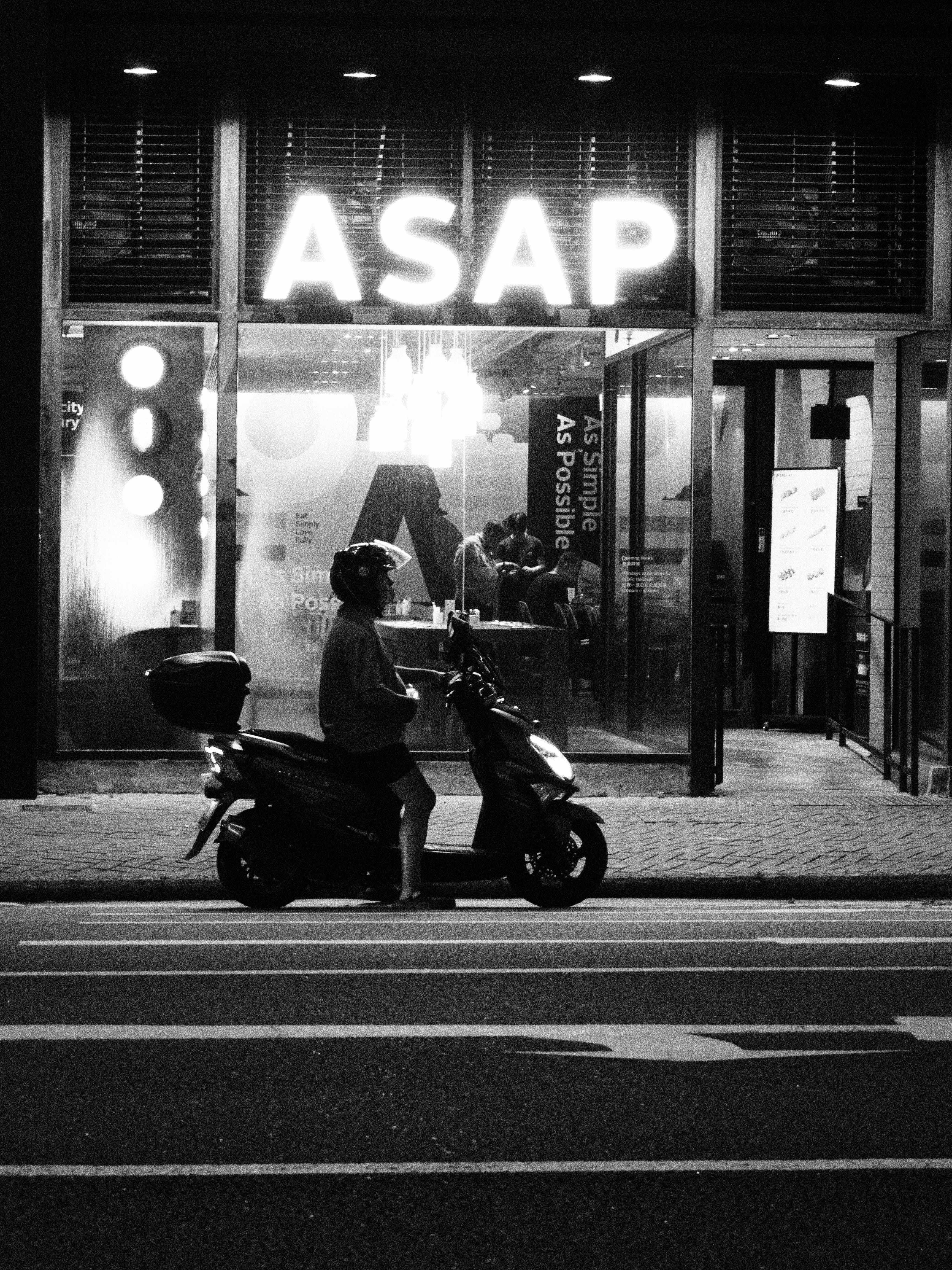 Black and white image of a person on a moped outside a Hong Kong street shop at night.