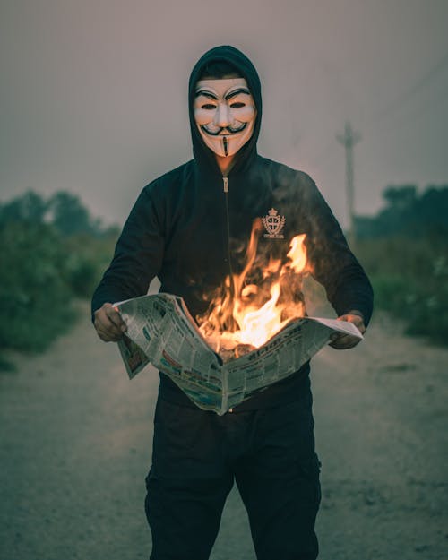 Person wearing Guy Fawkes Mask holding a Burning Newspaper