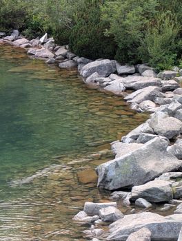 Tranquil rocky lakeside with clear water reflecting greenery, perfect for nature and travel themes.