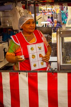 A female vendor serves food at a vibrant market stall, wearing a hairnet and apron.