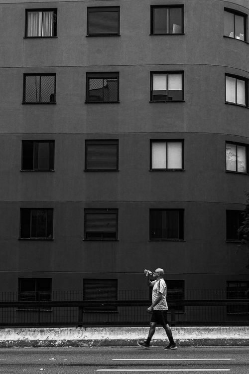 Free Black and white photo of a man walking past a modern apartment building, showcasing urban lifestyle. Stock Photo