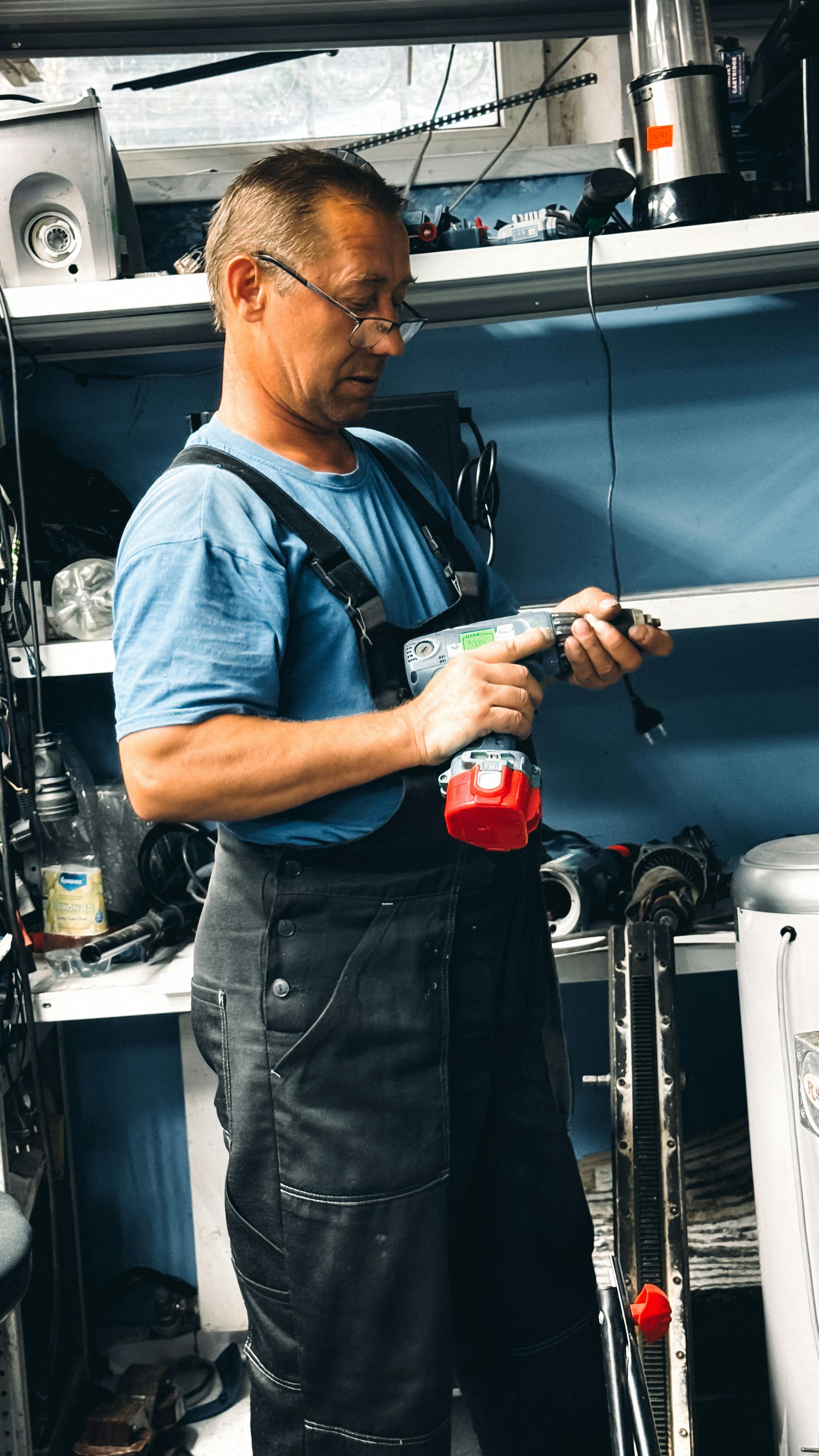Middle-aged man using a drill in a cluttered workshop setting.