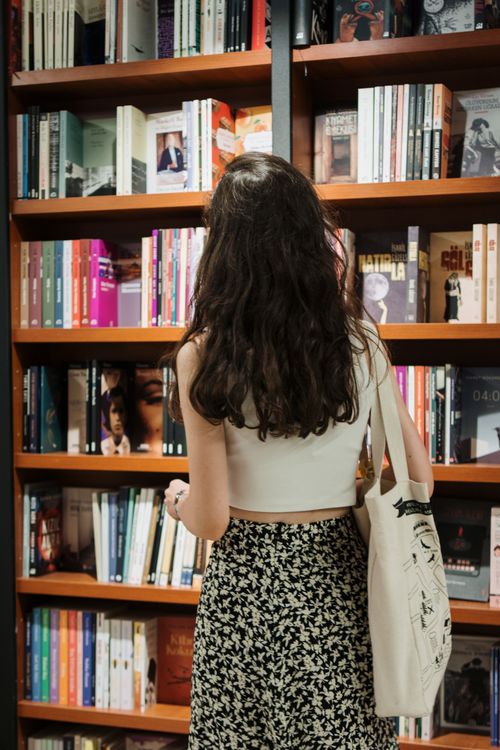 Free A woman with long hair looks at books on a shelf in a bookstore, carrying a canvas bag. Stock Photo