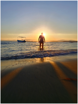 A person walks into the ocean at sunset creating a dramatic silhouette with a boat in the background.