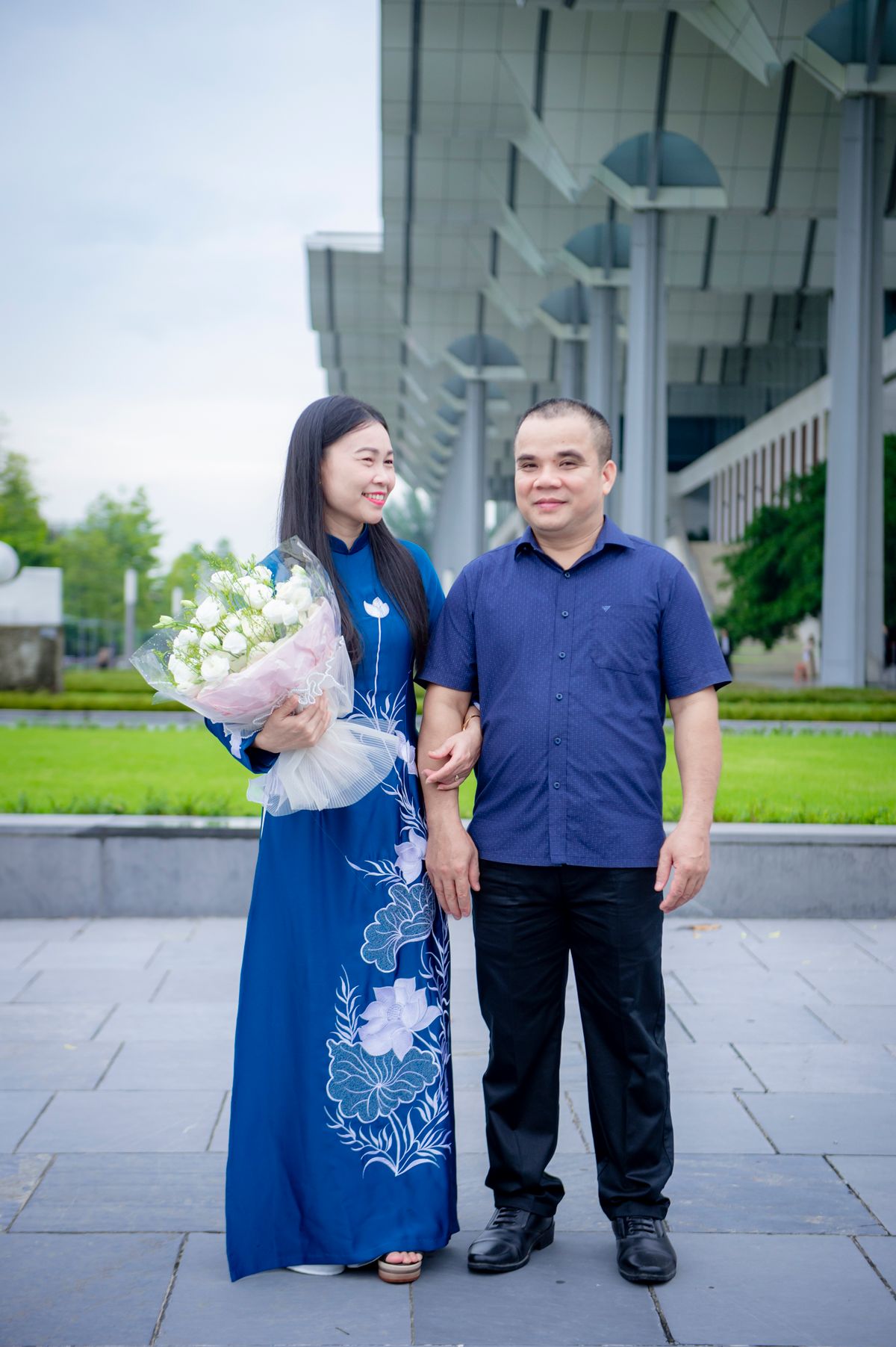 Cặp đôi trung niên khoác tay, người phụ nữ áo dài xanh ôm hoa trắng