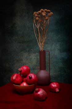 A classic still life featuring vibrant red pomegranates and a stylish tall vase against a textured backdrop.