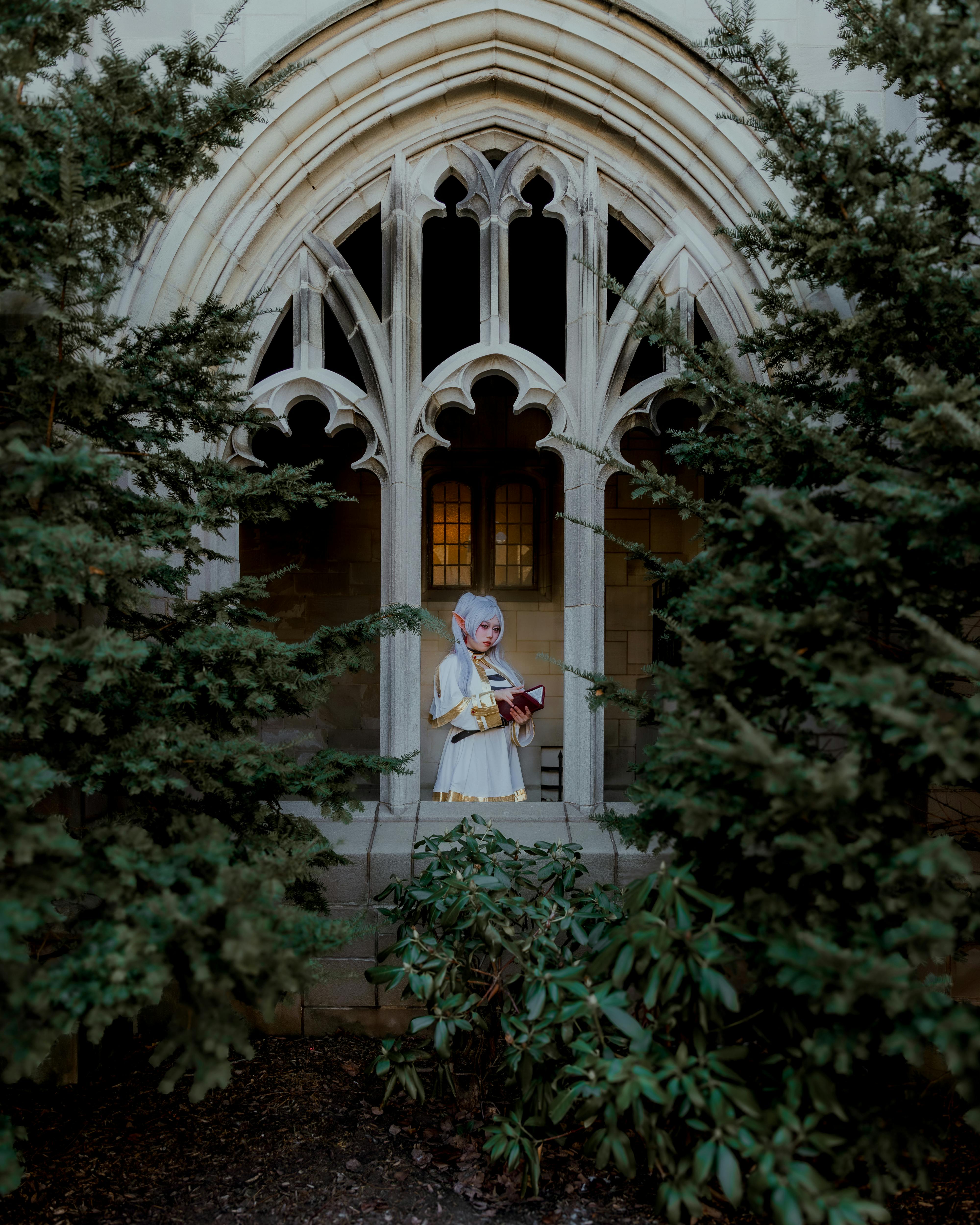 Cosplayer in white and gold costume stands within gothic architecture surrounded by greenery.