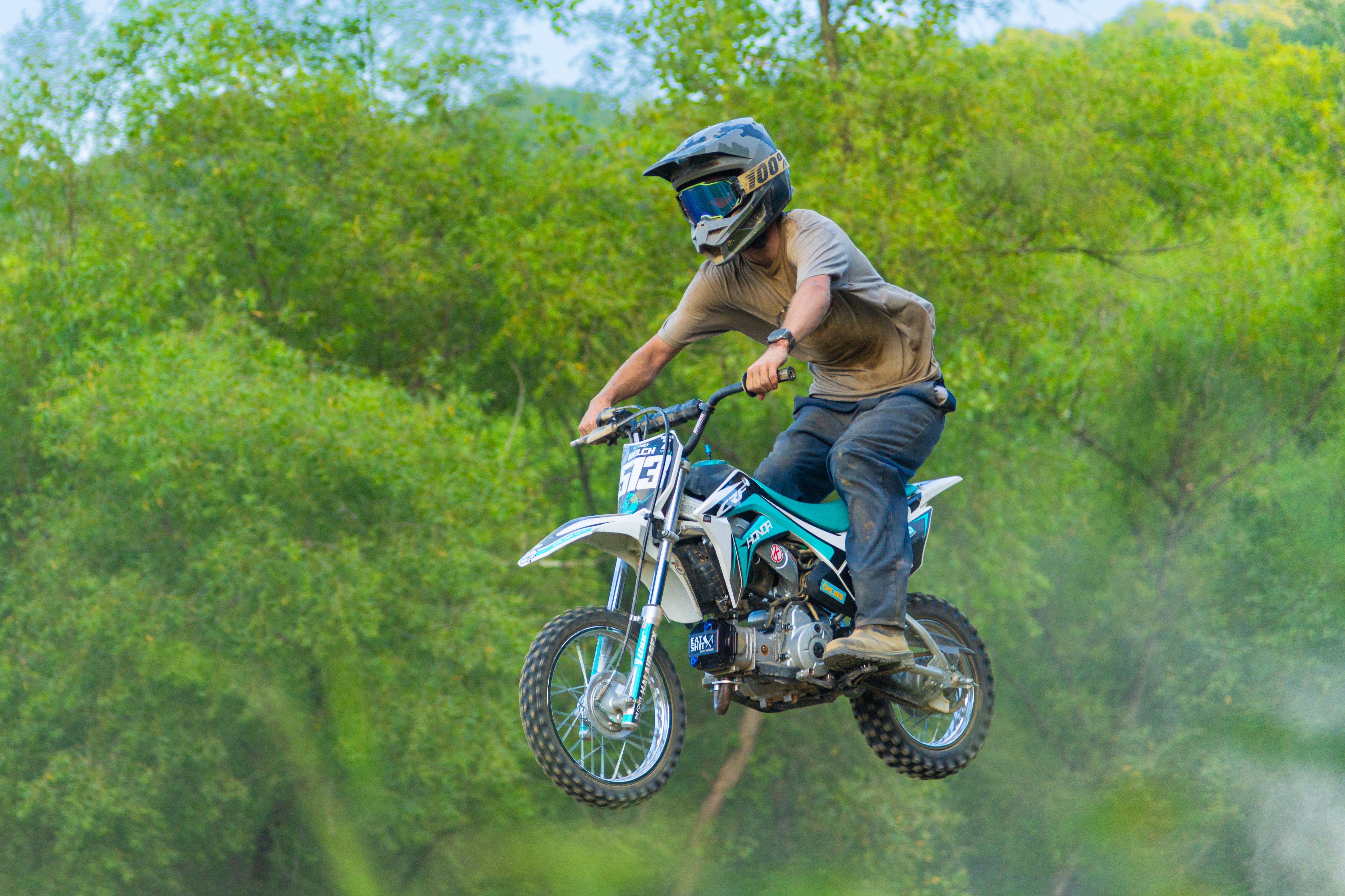 A motocross rider performs an impressive jump on a dirt bike track surrounded by lush greenery.