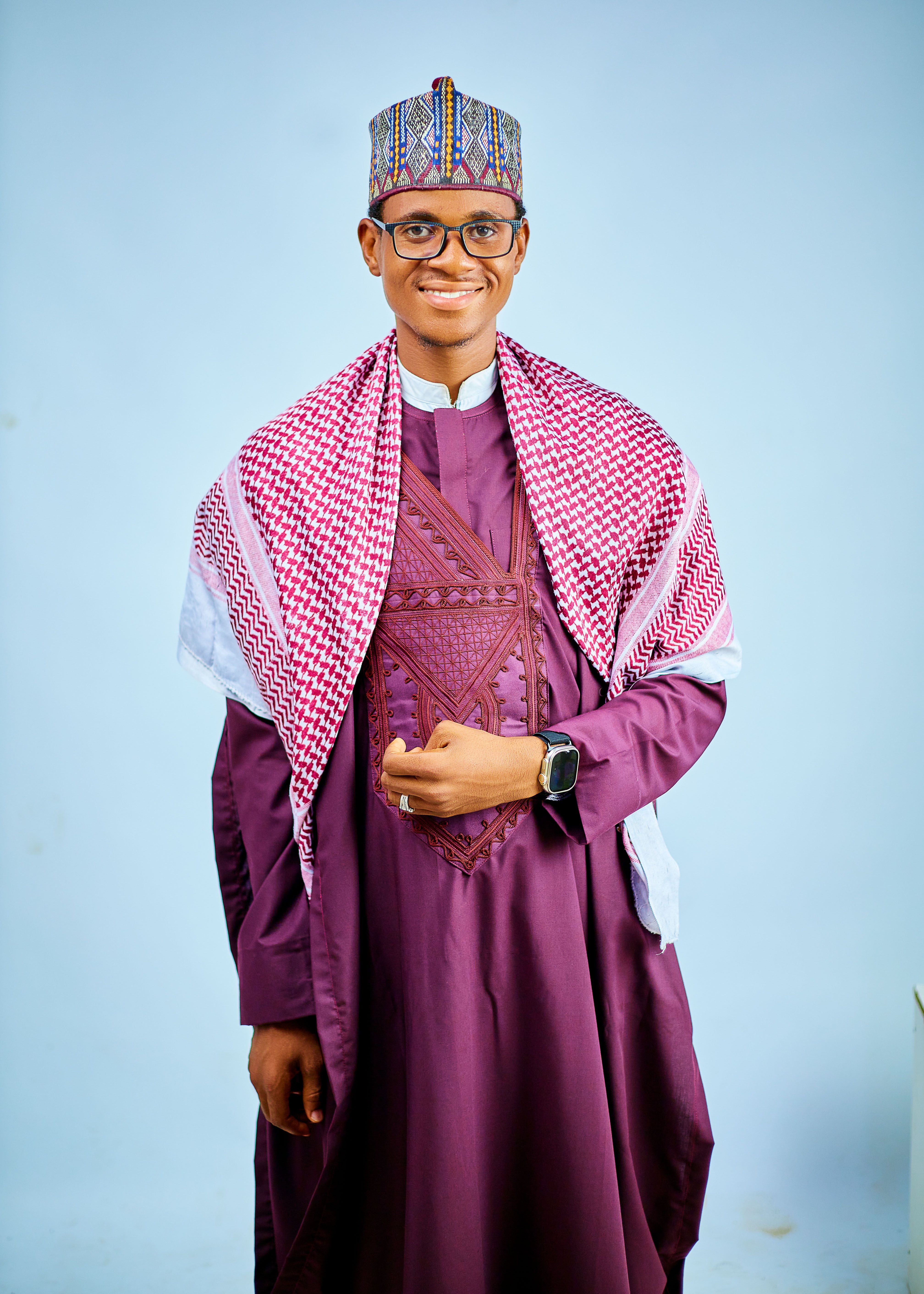 Smiling man in purple traditional attire with patterned headwear on a light background.